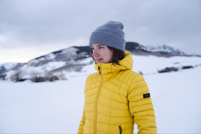 A woman wearing a bright yellow decathlon puffer jacket, standing in a snowy landscape with mountains in the background