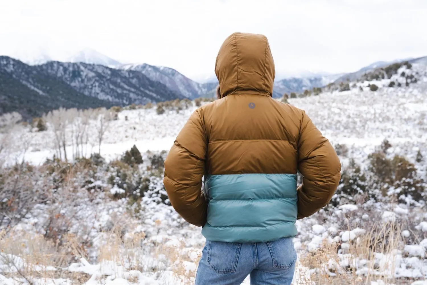 Person wearing marmot hoody stands in a snowy mountain landscape, looking at the distant snow-covered peaks