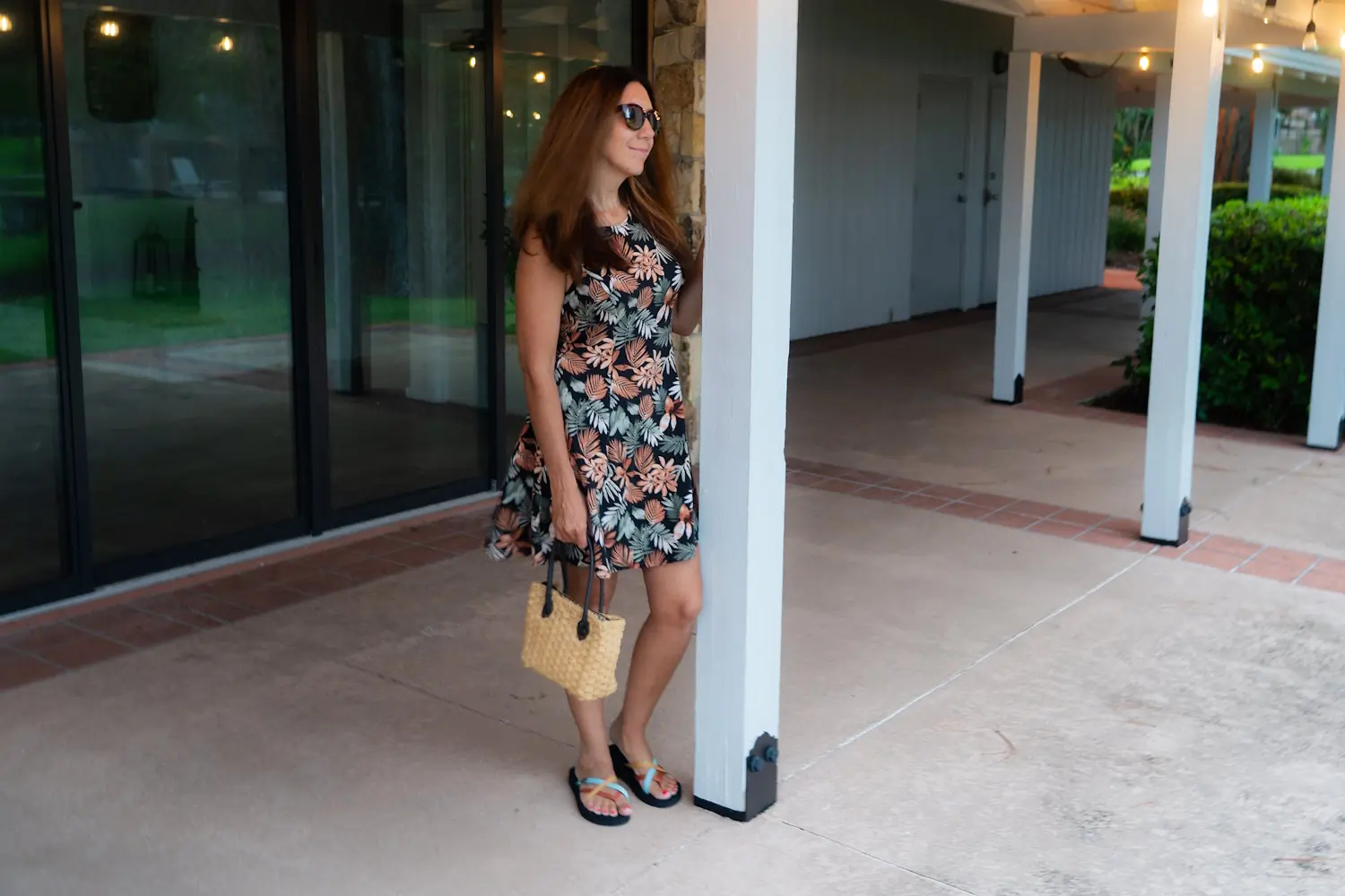 A woman in a floral dress and sunglasses holding a straw bag and wearing Teva Olowahu flip-flops