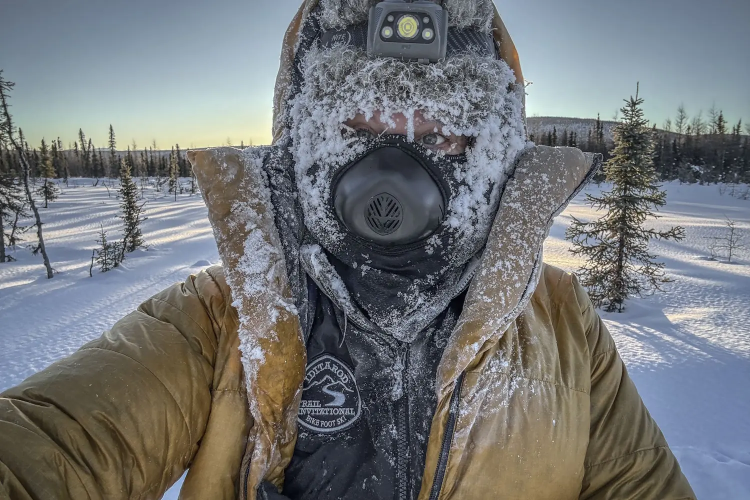 Person bundled up in heavy winter gear with a frost-covered face in a snowy landscape