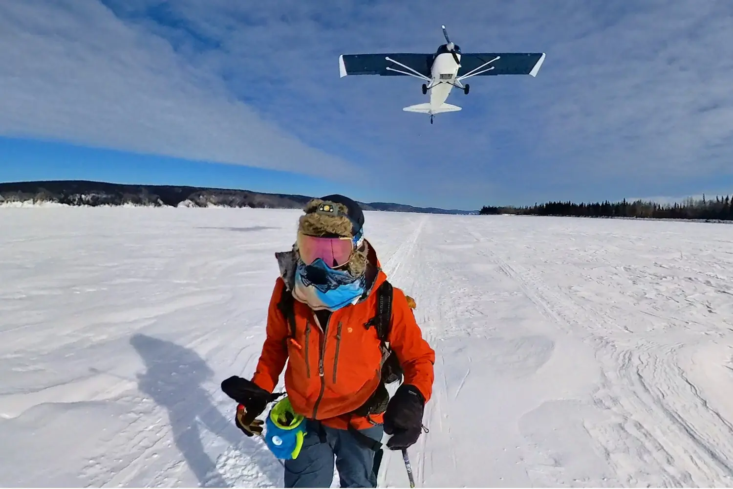 A person dressed in winter gear stands on a snowy landscape with a small airplane flying low overhead