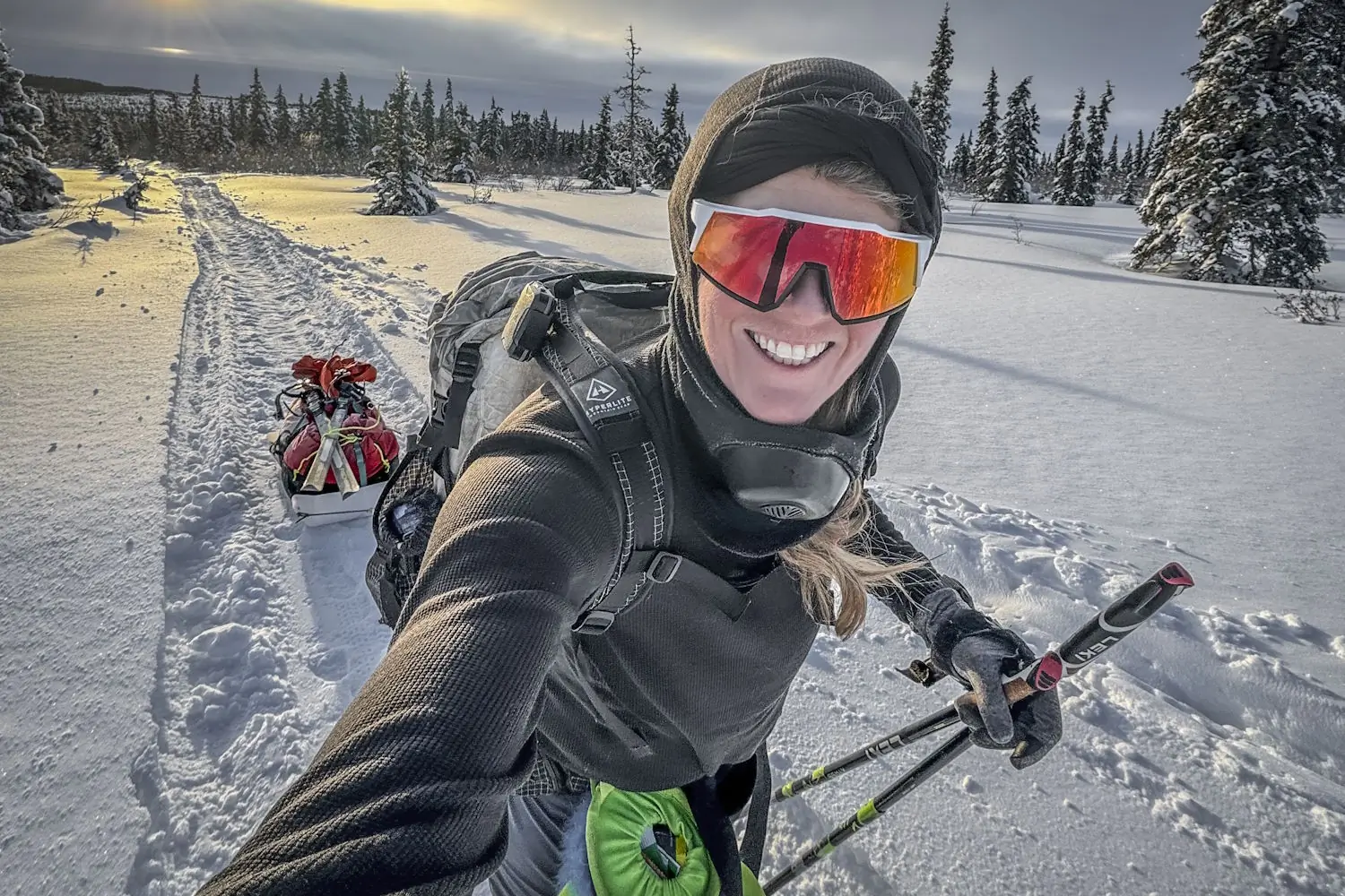Skier in winter gear pulling a sled through a snowy forest at sunset