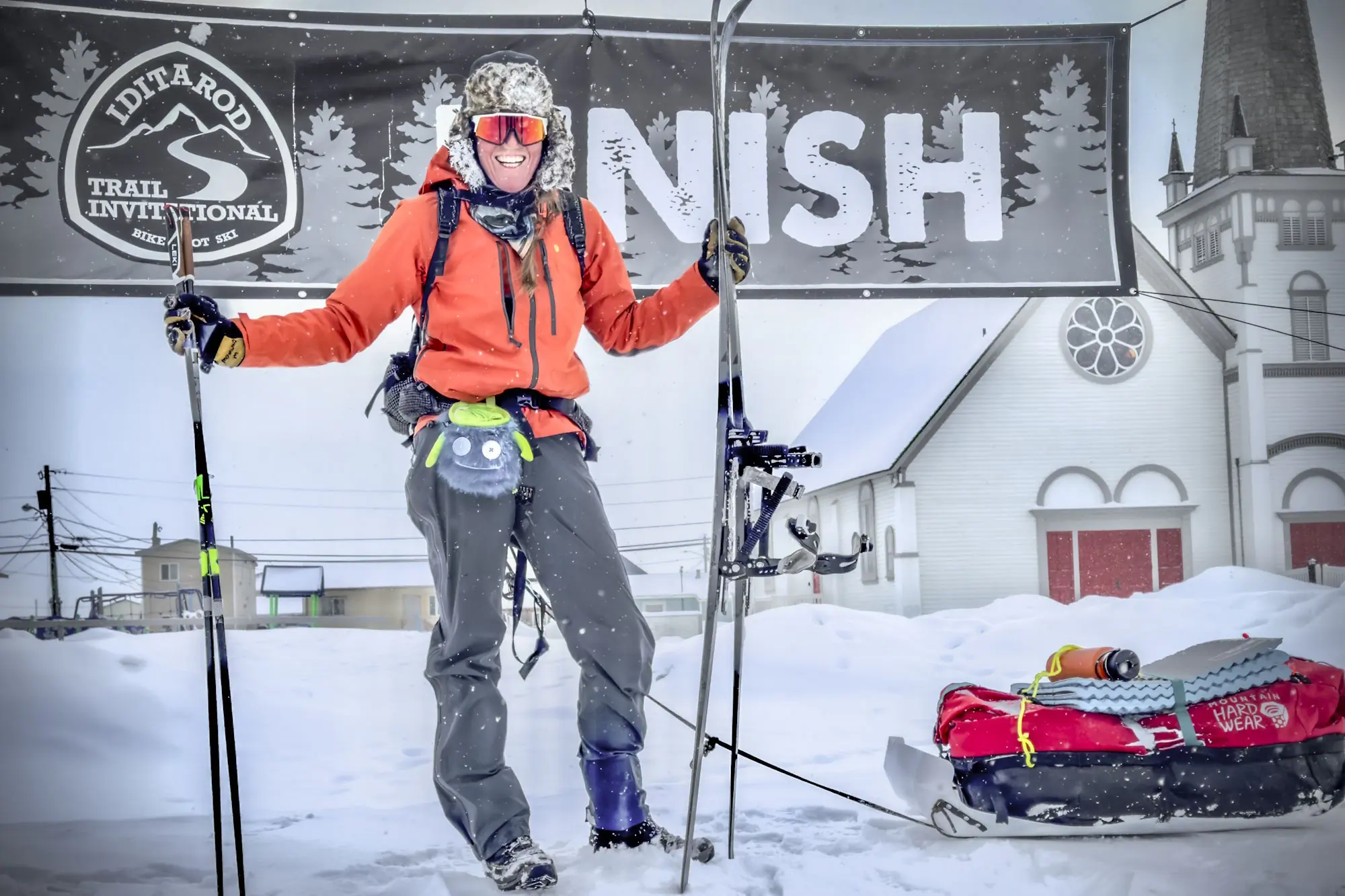 Smiling skier at the Iditarod Trail Invitational finish line, standing in snow with ski poles and gear