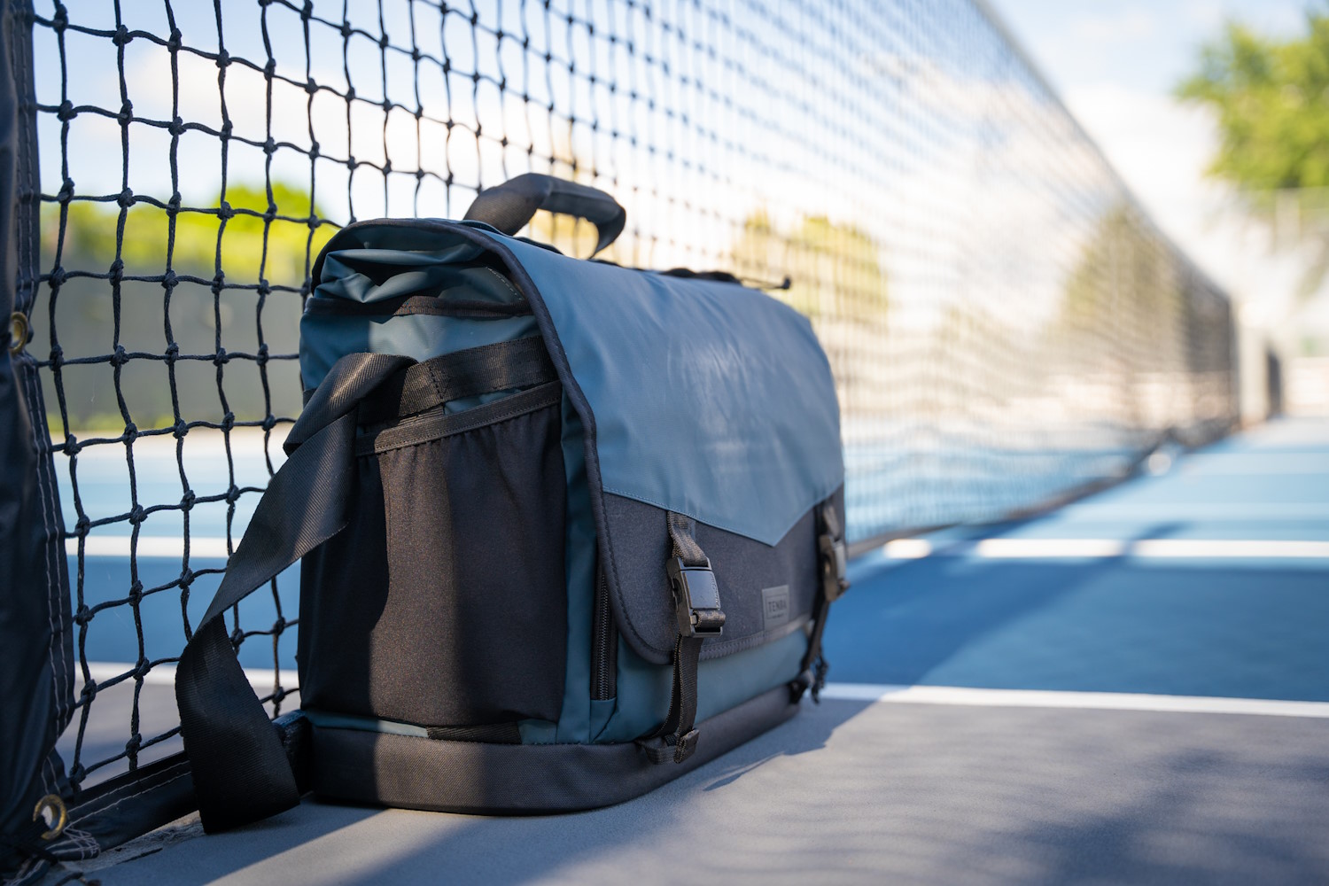 A Tenba DNA 16 Pro Messenger Bag resting against a tennis court net
