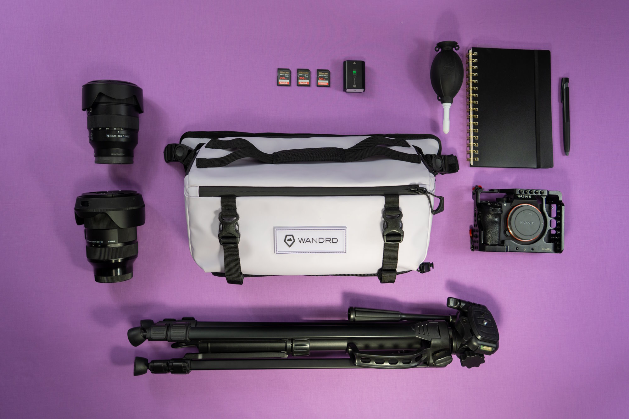 A flat lay of photography gear, including a WANDRD camera bag, lenses, a Sony camera, tripod, and accessories on a purple background