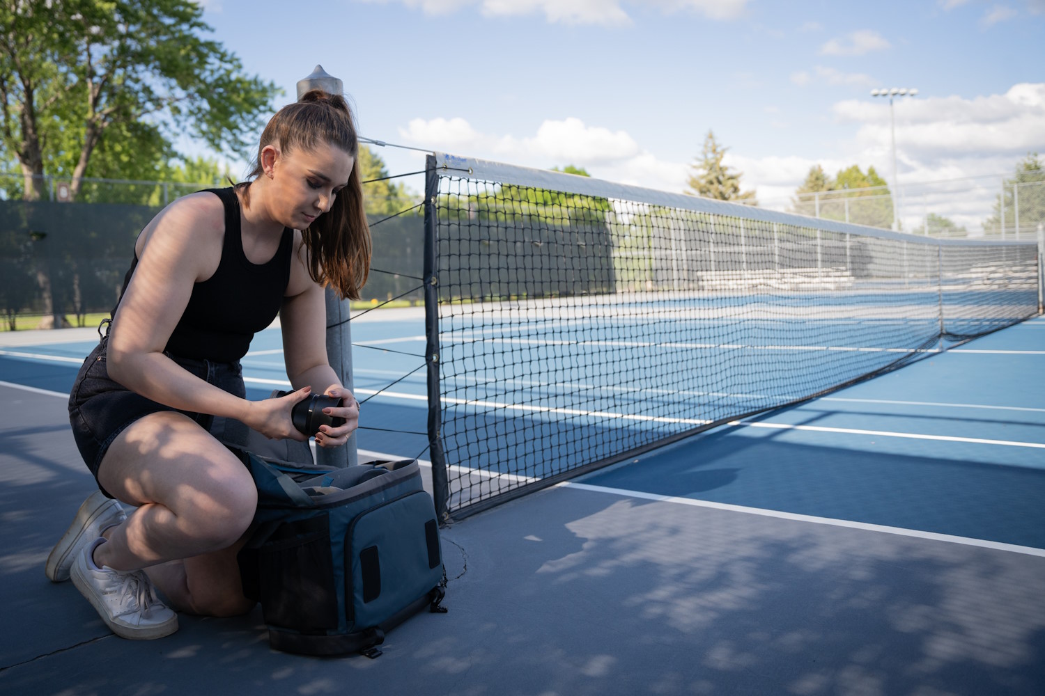 A woman kneels by a tennis court, placing a camera lens into a blue Tenba DNA 16 Pro bag