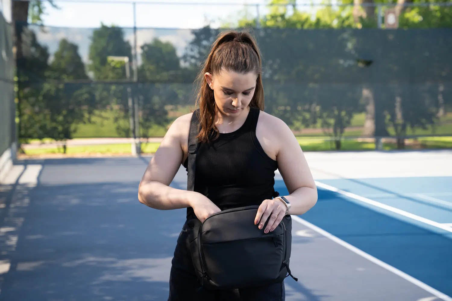 A woman standing on a tennis court, focused on opening a black crossbody bag