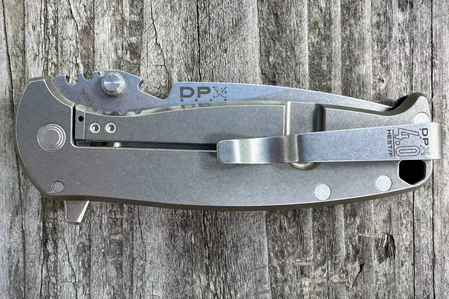A folded DPx Gear HEST/F 4.0 knife with a titanium clip rests on a textured wooden surface
