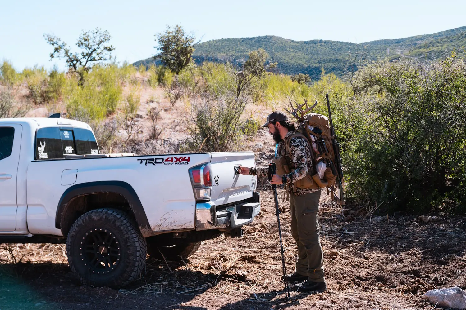 Hunter standing beside a white Toyota TRD 4x4 truck with gear and a rifle in a desert