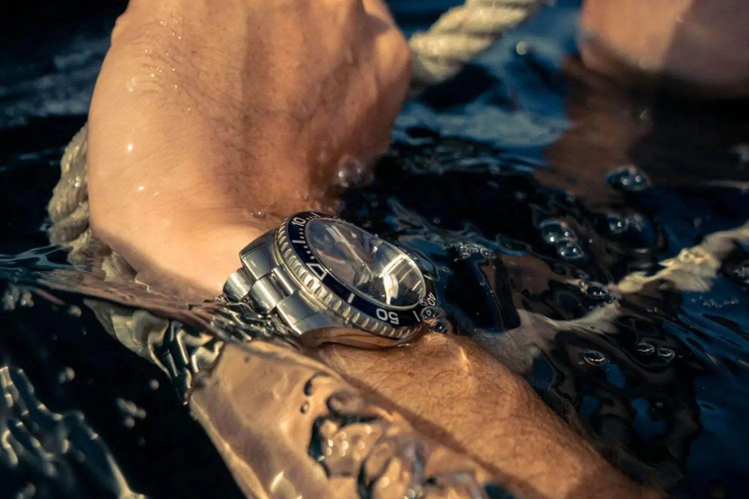 A wrist wearing a stainless steel dive watch submerged in water while gripping a rope