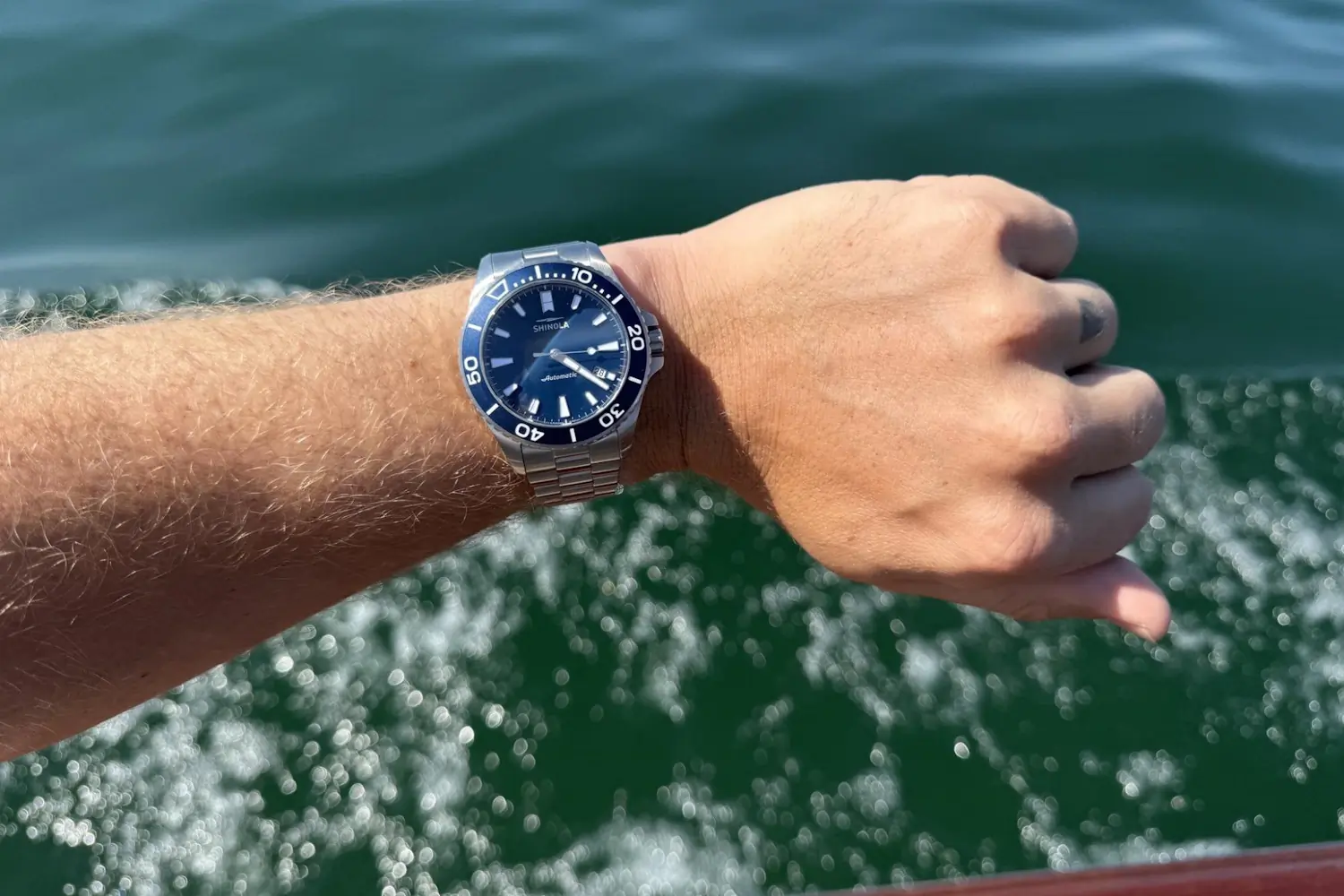 Michigan Monster watch with a blue dial is worn on a wrist held out over the water