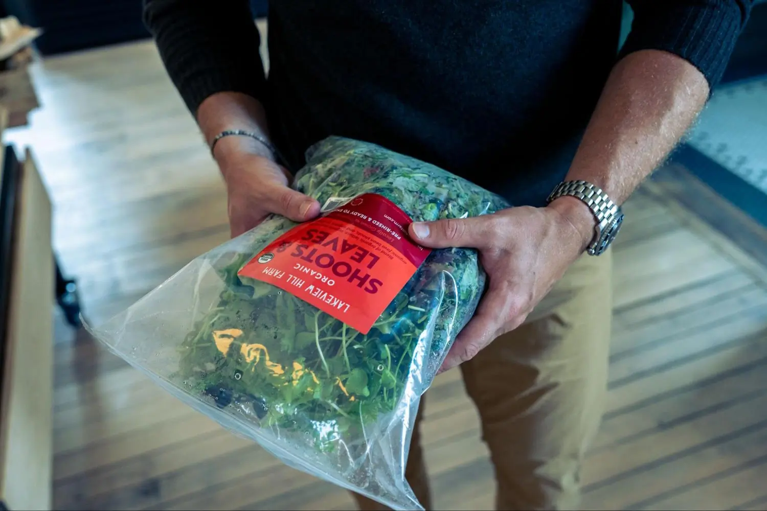 A person holds a bag of organic greens labeled Shoots & Leaves