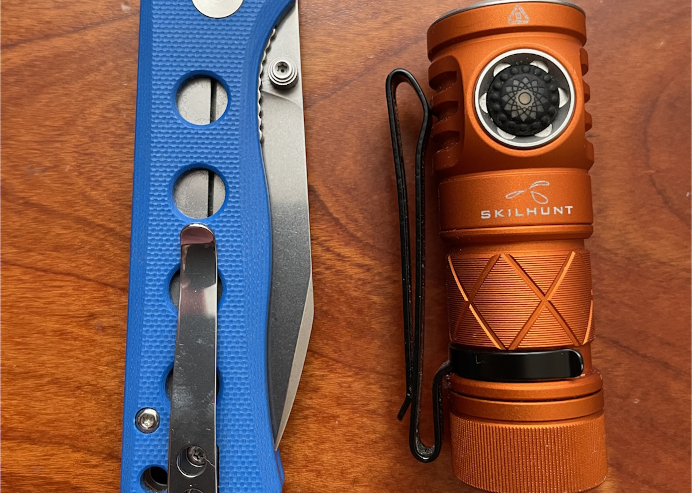 A folded canary knife next to an orange Skilhunt flashlight on a wooden surface