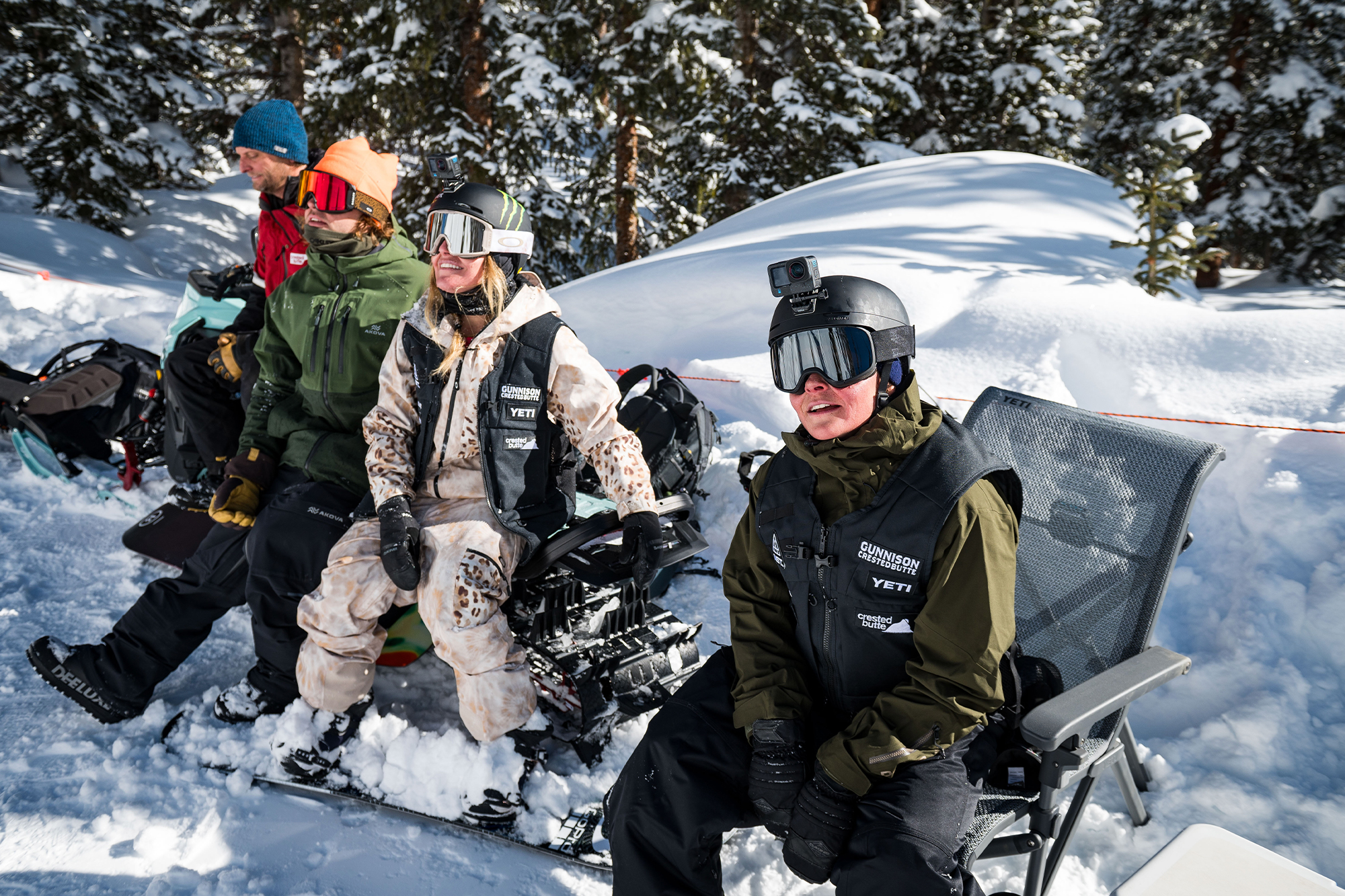 Snowboarders sit in the snow, geared up in helmets, goggles, and winter jackets, watching the action at the Natural Selection event