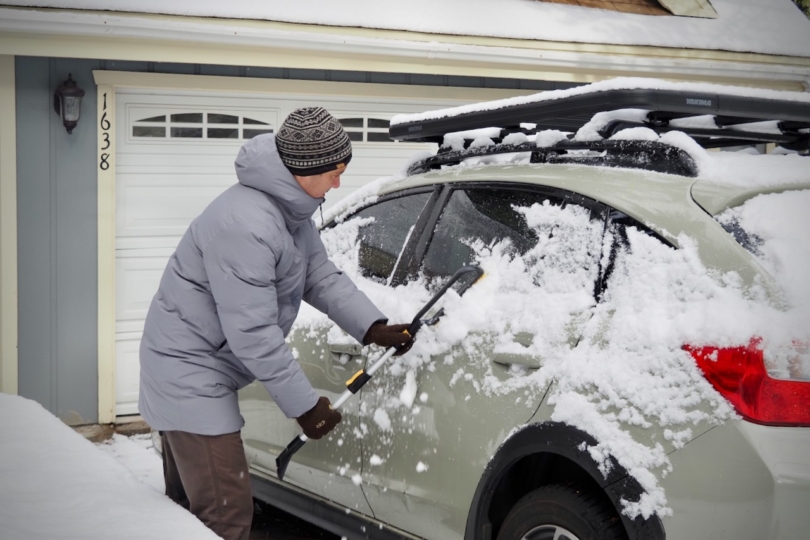 A man is using a snow scraper to brush snow off of a Subaru Crosstrek, while wearing the Patagonia Jackson Glacier Parka.