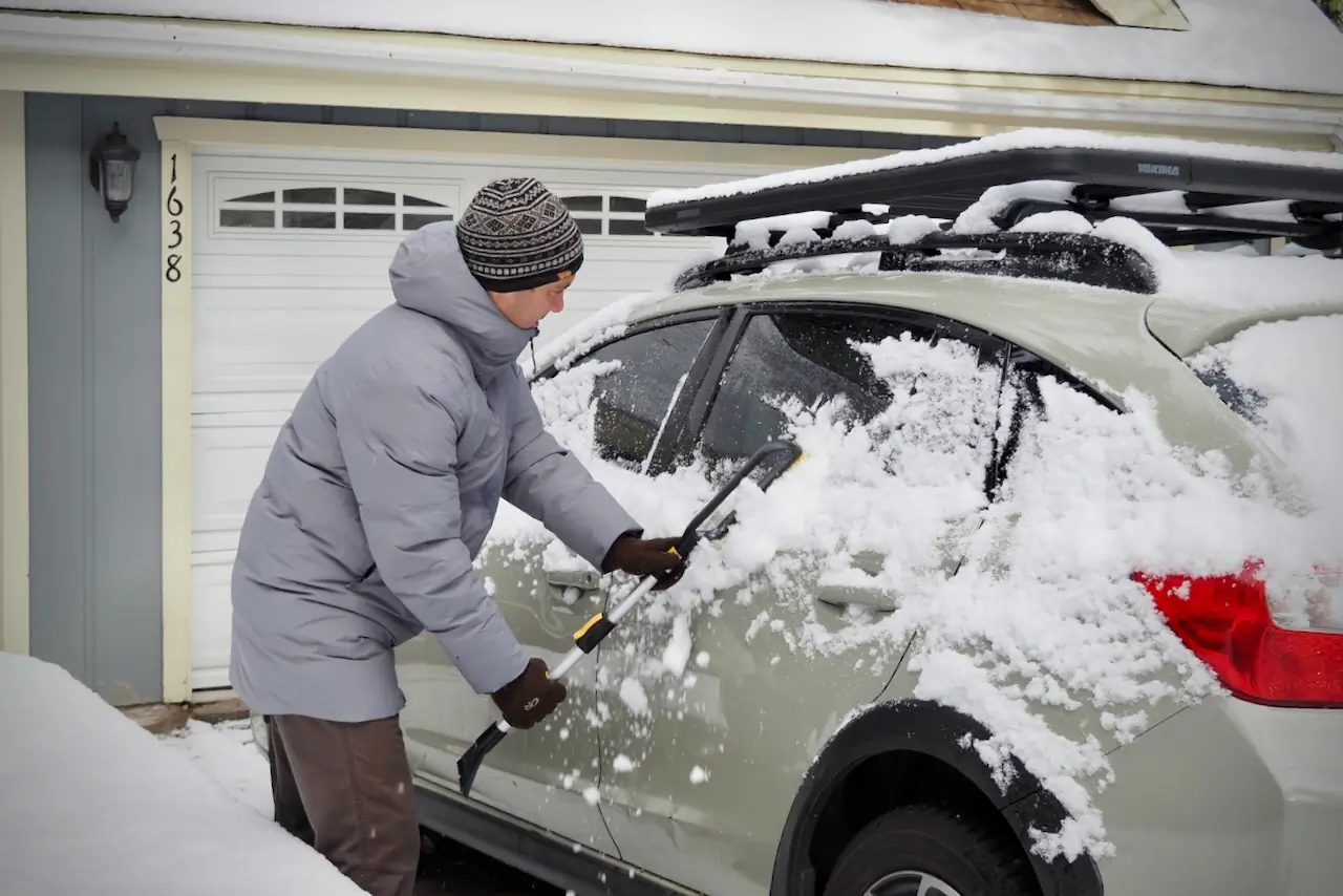 A man is using a snow scraper to brush snow off of a Subaru Crosstrek, while wearing the Patagonia Jackson Glacier Parka.