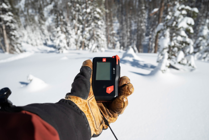 A gloved hand holds an avalanche beacon in a snowy forest clearing