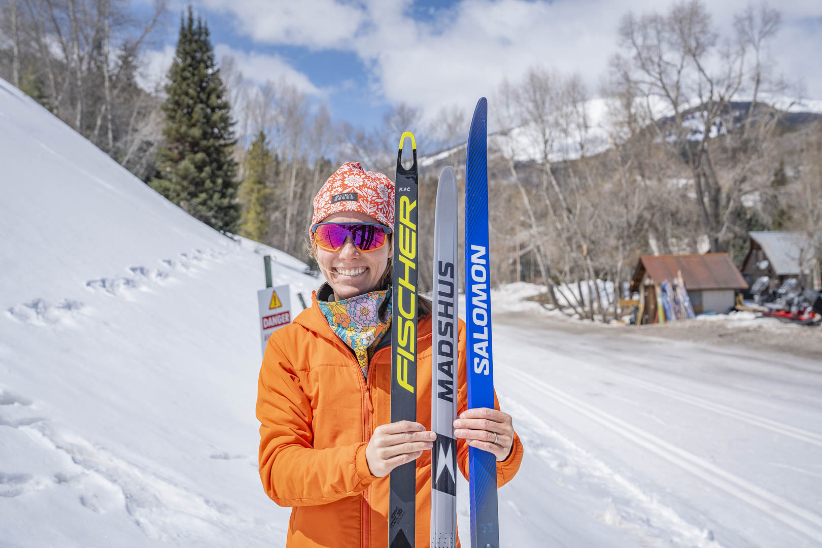 Smiling person holds three skate skis on a snowy trail with a forested mountain backdrop