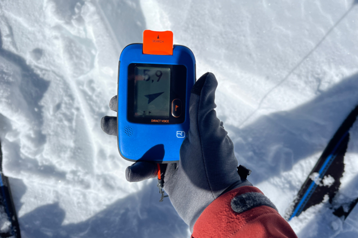 Gloved hand holding an avalanche beacon displaying search mode in snowy terrain