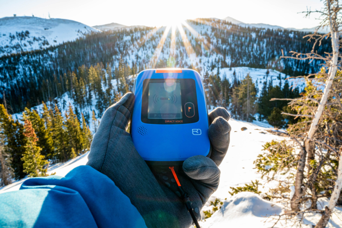 Gloved hand holding Ortovox Diract Voice beacon with sunlit snowy mountains in the background