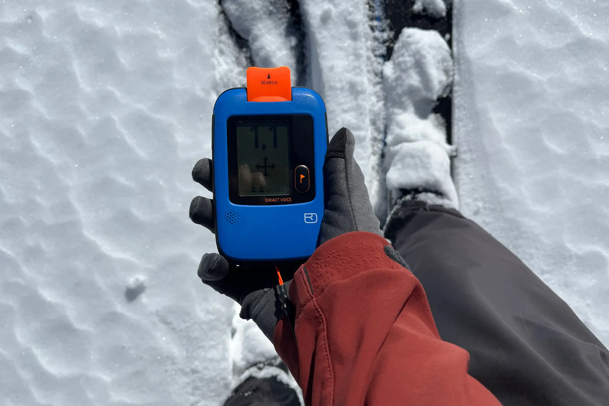 Gloved hand holding an avalanche beacon in search mode over snowy tracks