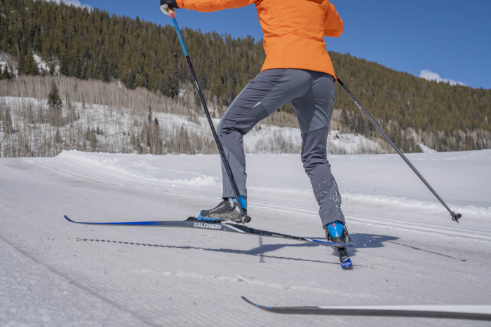 Skier wearing orange jacket glides on a snowy trail surrounded by forest.