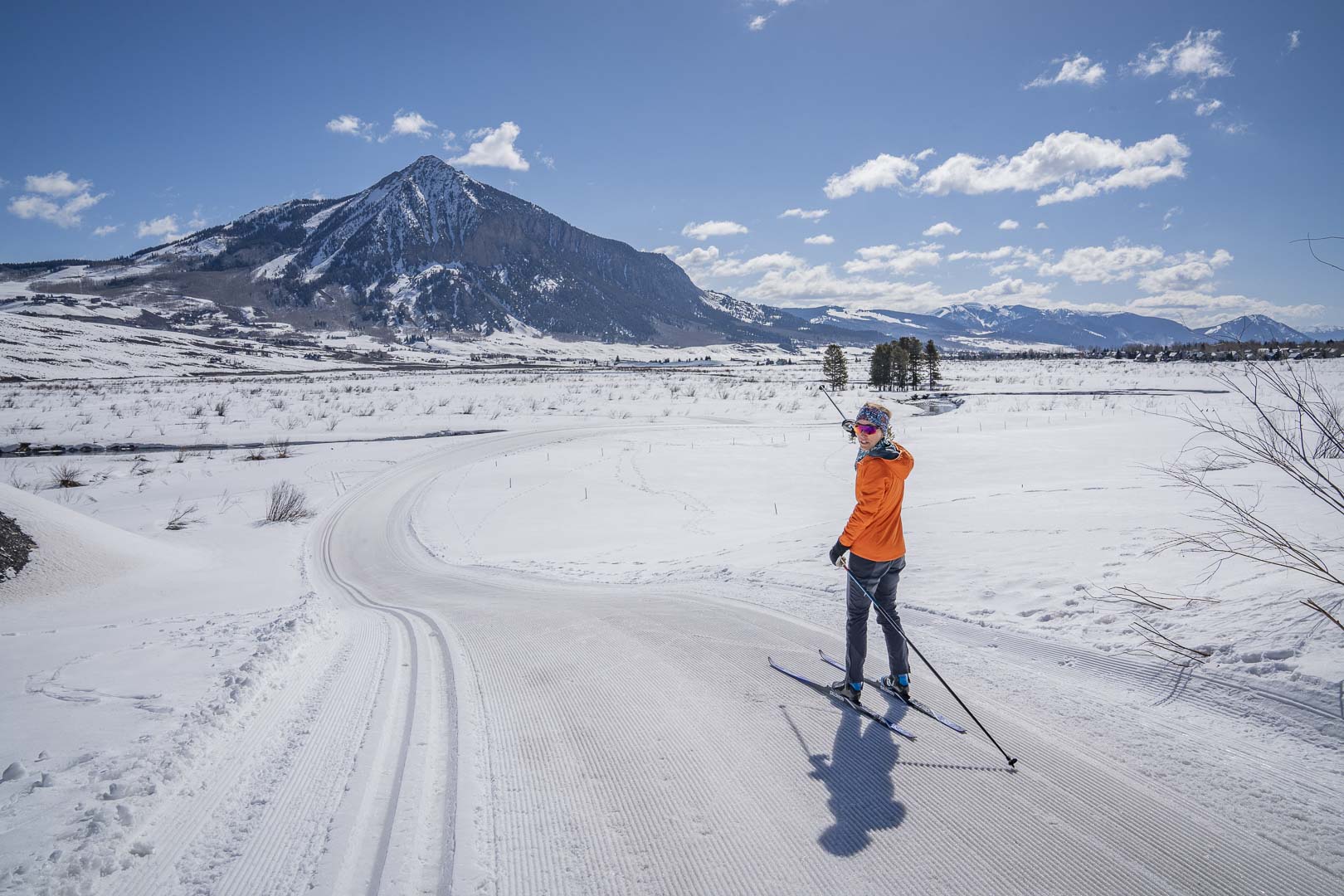 Two skiers pose and smile on a groomed trail with snowy mountains behind them