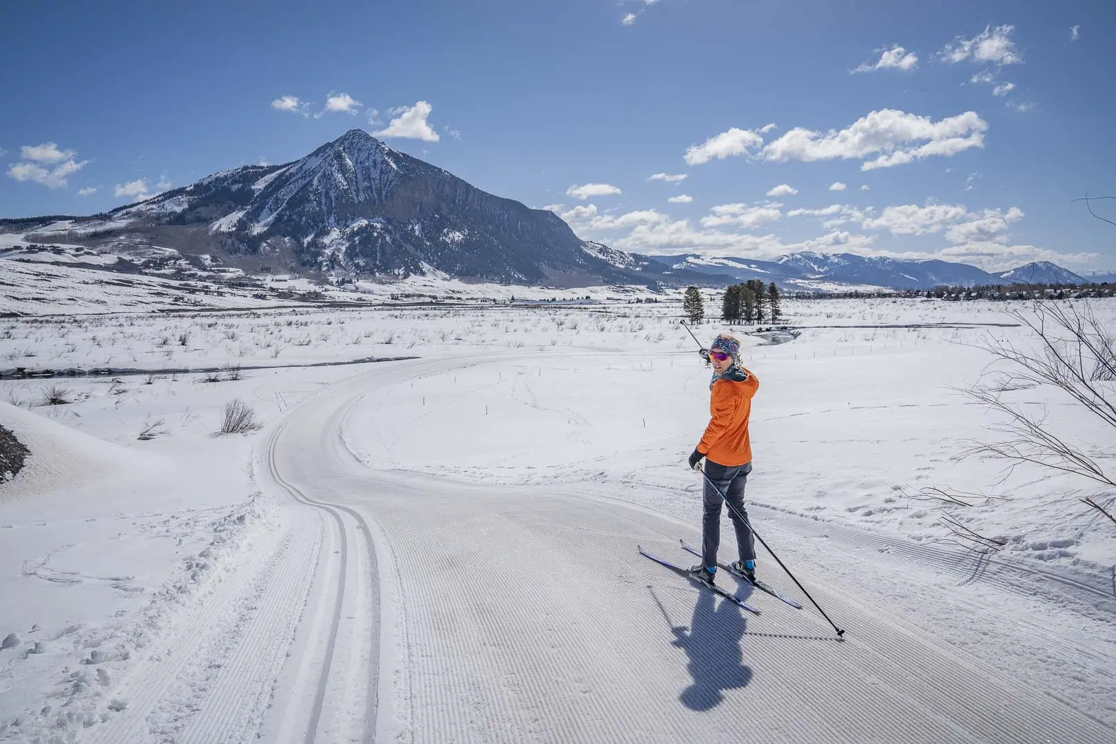 Two skiers pose and smile on a groomed trail with snowy mountains behind them