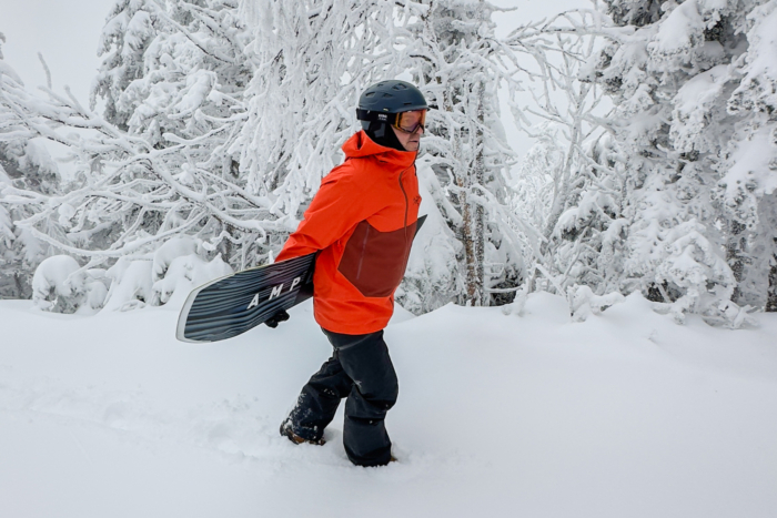 Person walking through deep snow, carrying a snowboard, surrounded by snow-covered trees