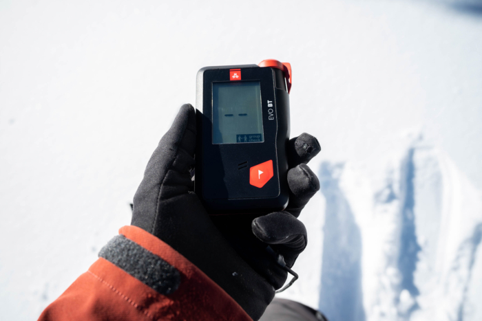 A gloved hand holds an avalanche beacon in group check mode on snowy terrain