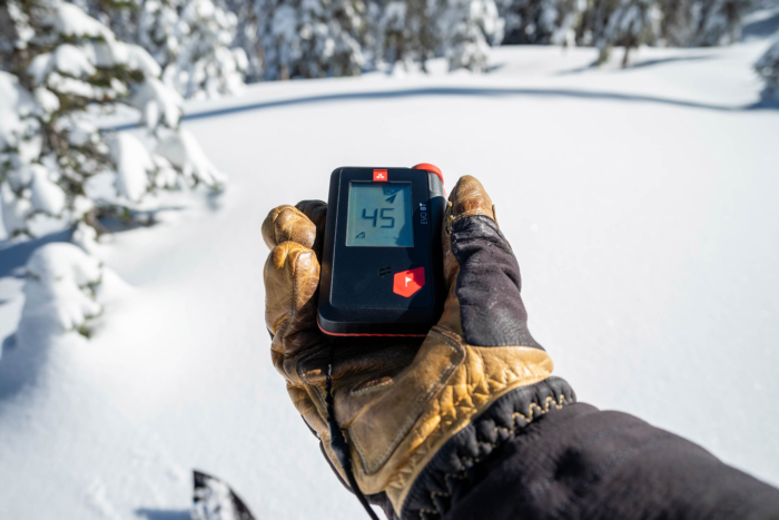 A gloved hand displays an avalanche beacon reading '45' in a snowy forest