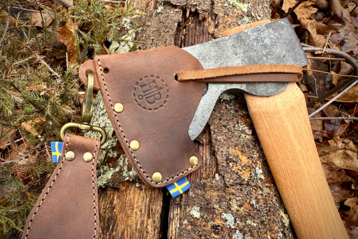 Close-up of the Hults Bruk Spika axe with a leather sheath and Swedish flag tag on a mossy log