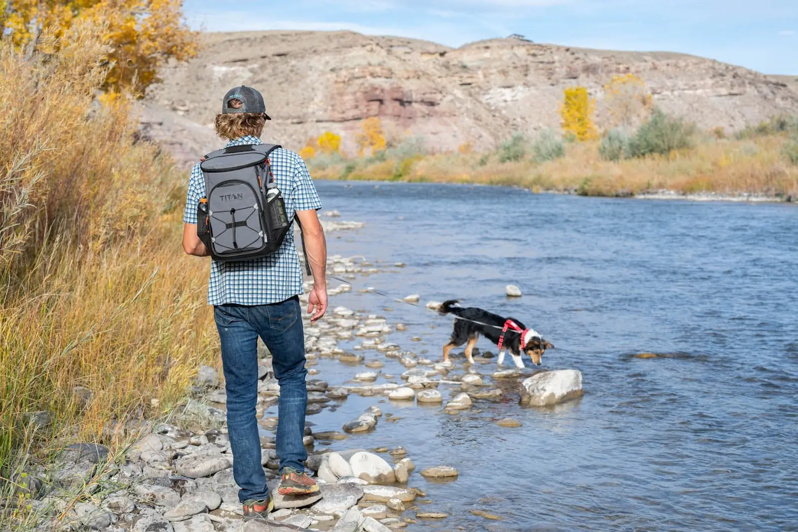 A man walks by a water with his dog, wearing a Titan Arctic Zone cooler backpack
