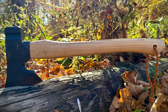 The Hardcore Hammers Camp Companion Axe lies on a log, with its wooden handle and braided lanyard against autumn leaves