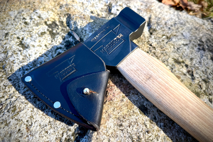 The Hardcore Hammers Camp Companion Axe with its leather sheath secured, resting on a textured rock surface under bright sunlight