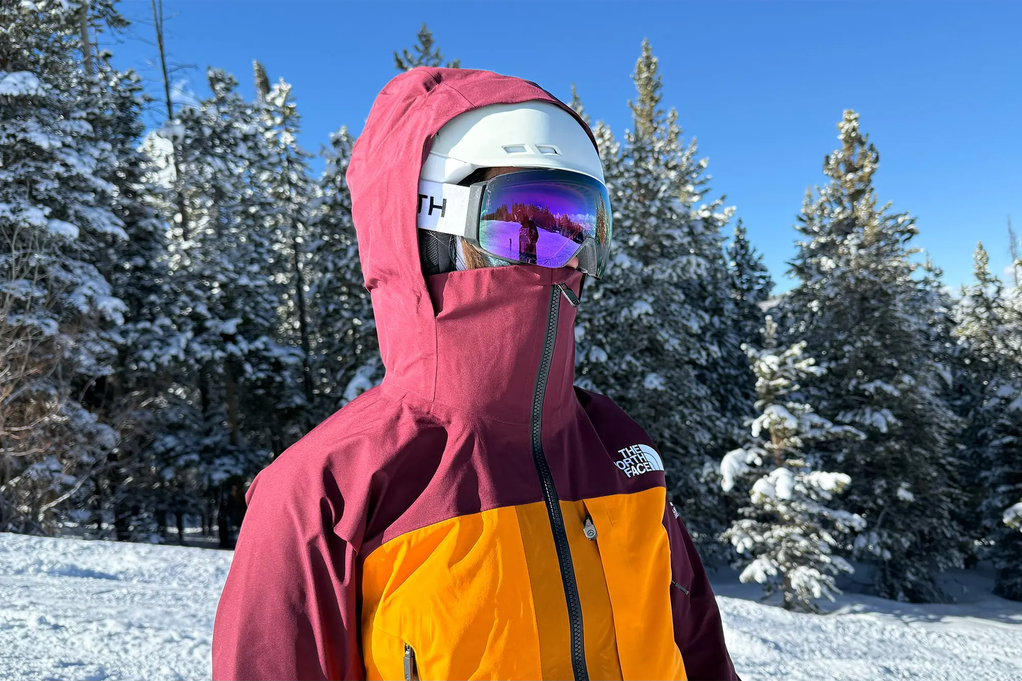 A close-up of The North Face Summit Series Verbier Gore-Tex Jacket showcasing its helmet-compatible hood in use, paired with a white helmet and reflective goggles against a snowy forest backdrop.







