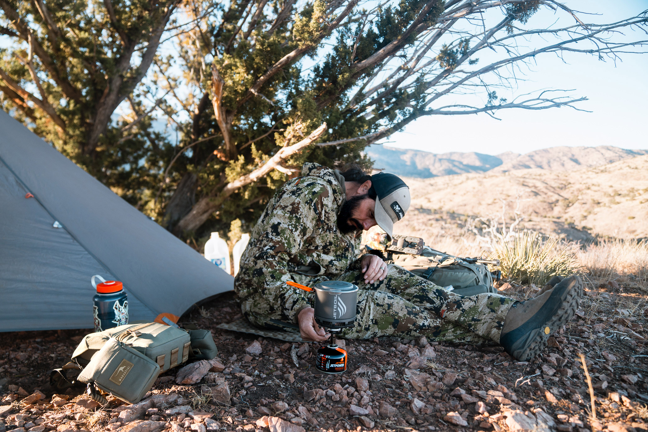 A camper in camouflage gear uses the Jetboil Stash Stove near a tent, with packs and water bottles nearby in a dry, mountainous landscape