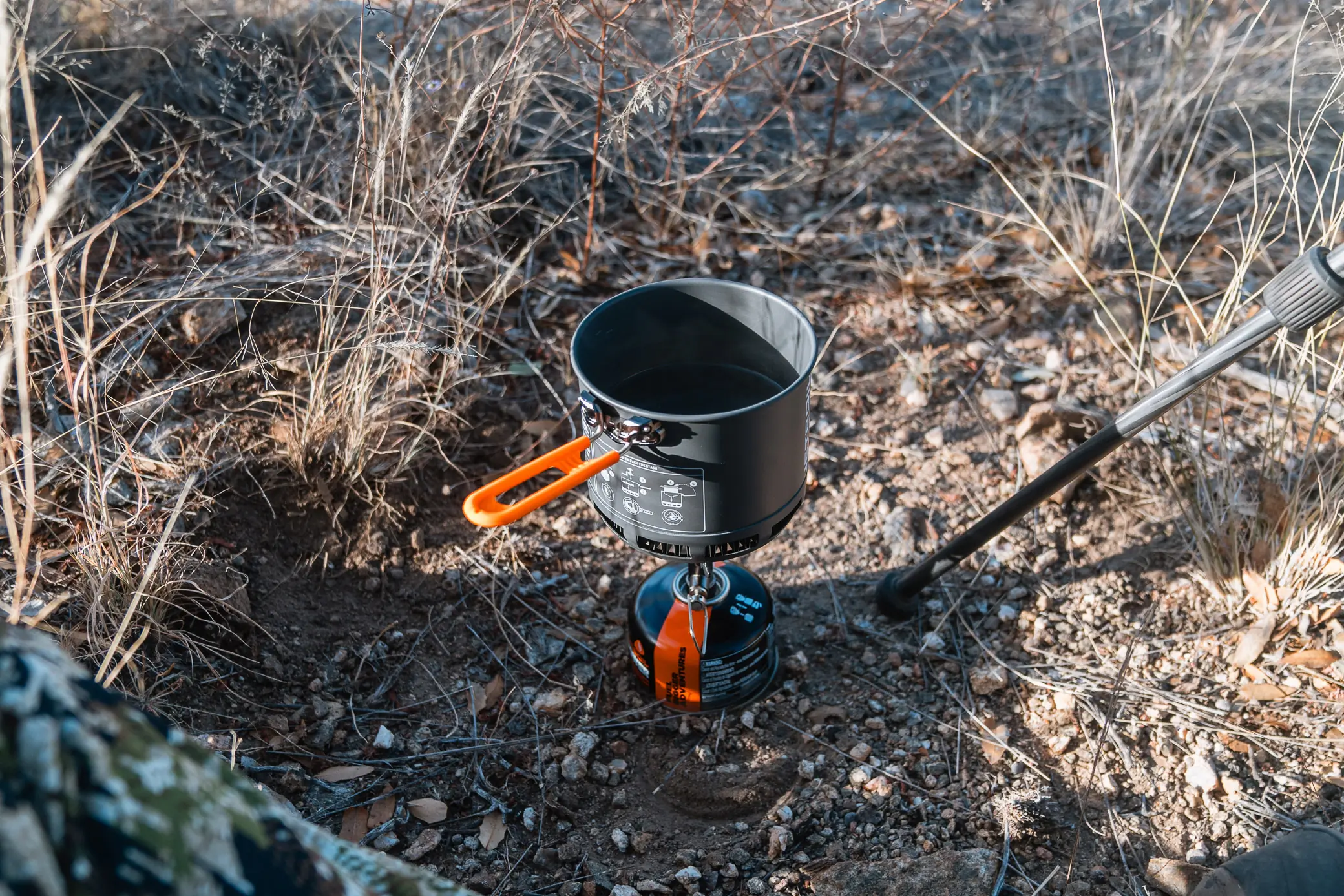 The Jetboil Stash stove set up on rocky ground, heating water in the pot with an extended orange handle