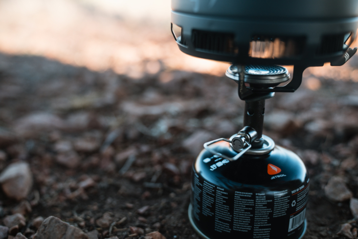 A close-up of the Jetboil Stash stove connected to a fuel canister, showing the burner and pot support in use on rocky ground