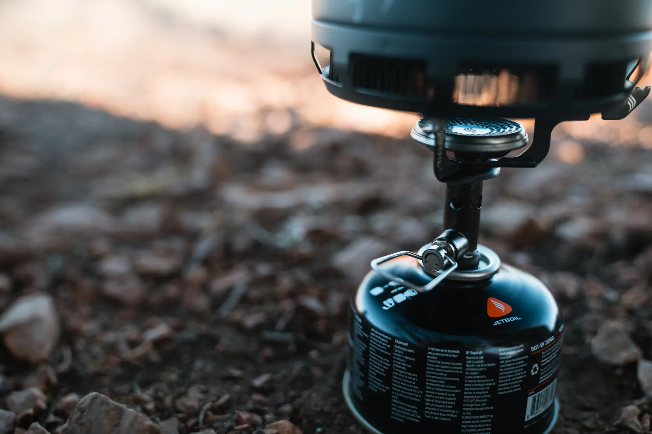 A close-up of the Jetboil Stash stove connected to a fuel canister, showing the burner and pot support in use on rocky ground