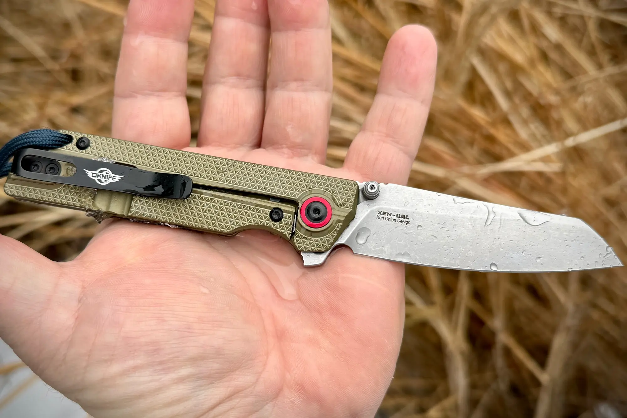 The open OKNIFE XEN-OAL knife resting in an outstretched hand, showing its size and detailing
