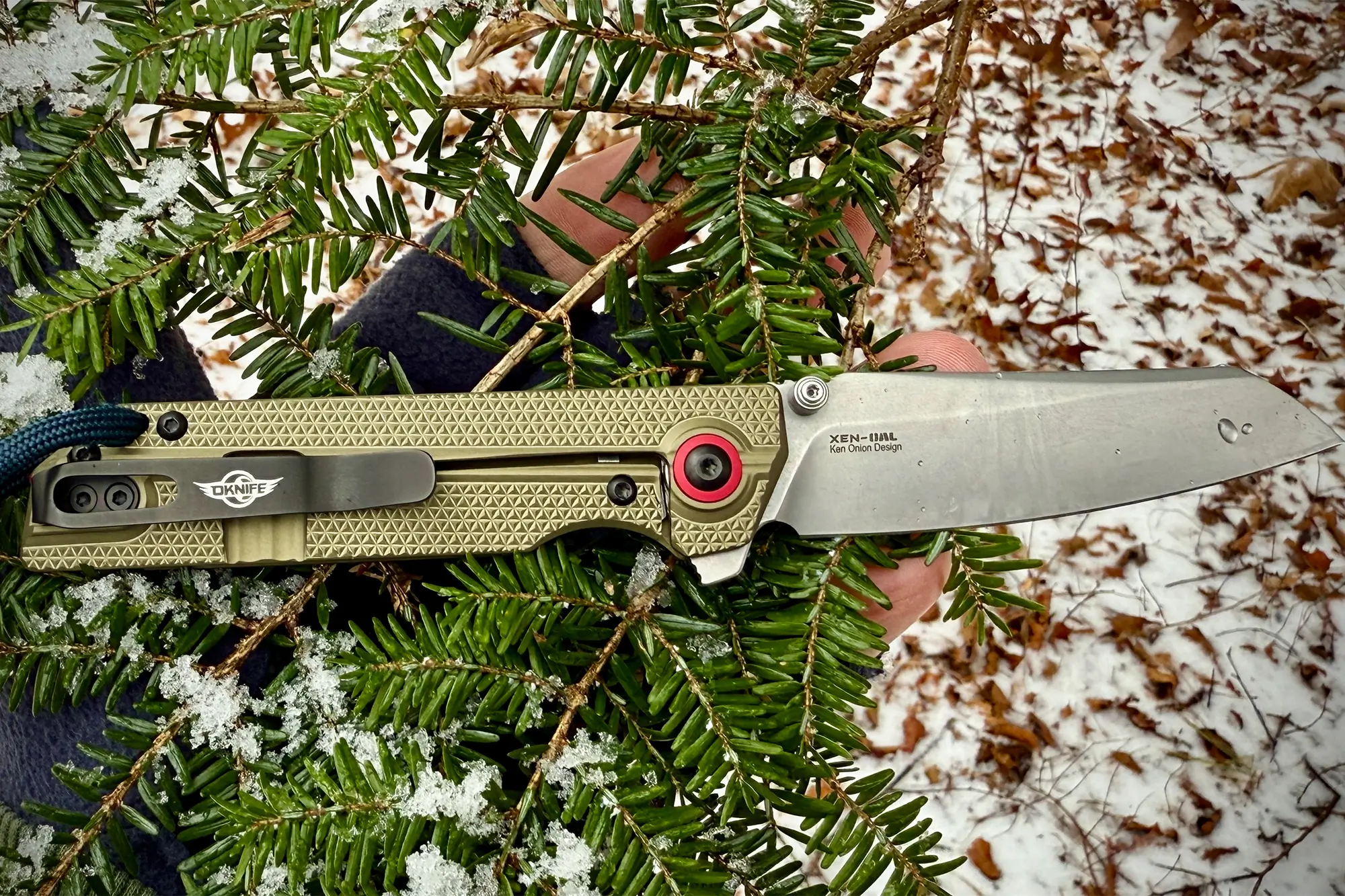 The open knife held among snow-dusted green fir branches, highlighting the olive green handle and silver blade
