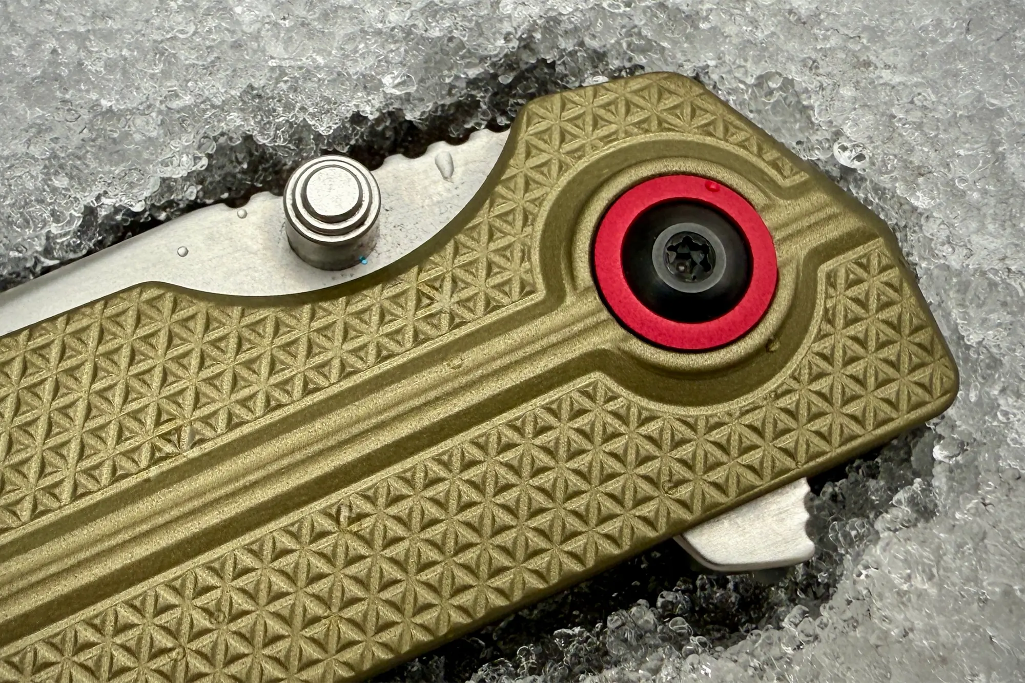 A close-up of the textured handle with a red pivot ring and silver thumb stud resting on icy snow