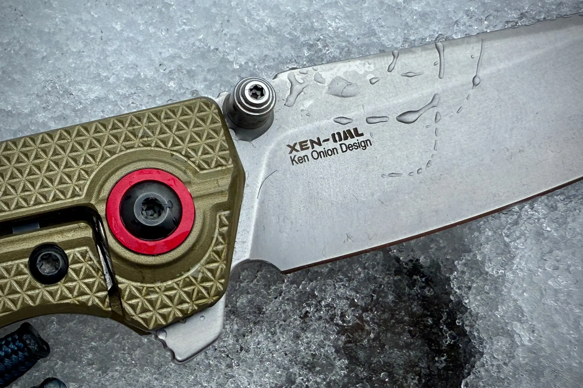  A close-up of the knife blade showing the "XEN-OAL" logo, "Ken Onion Design," and part of the pivot with a red accent