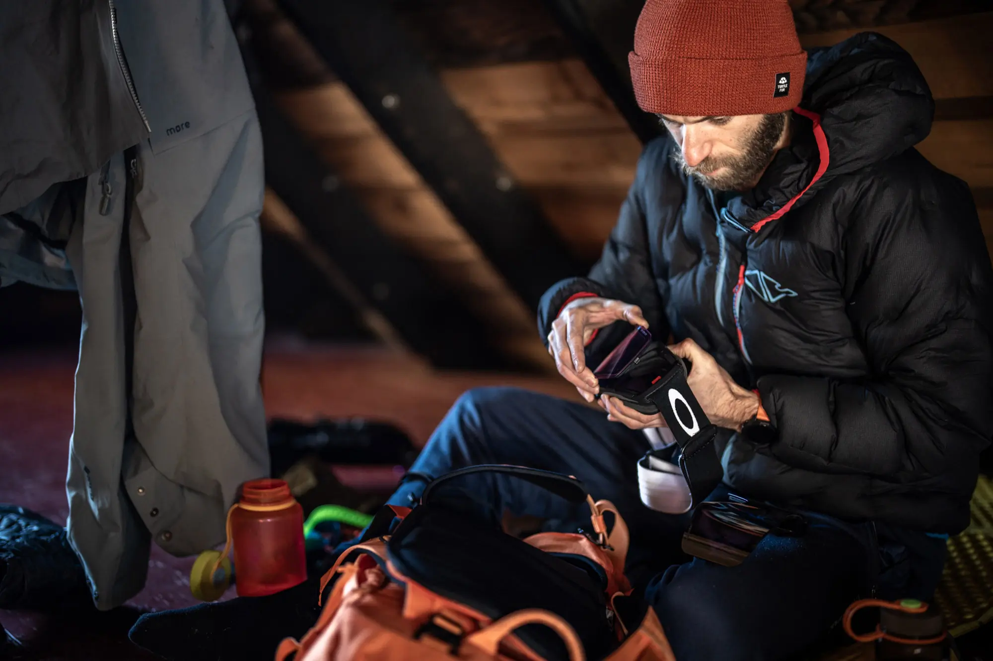 Inside a rustic cabin, a snowboarder is preparing the Oakley Line Miner Pro goggles, seated with gear scattered around and warm lighting creating a cozy atmosphere