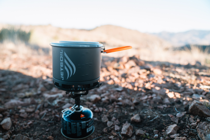 The Jetboil Stash Stove set up on rocky ground with a fuel canister, with a blurred mountainous landscape in the background