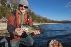 A smiling angler holds a bass with one hand, standing on a boat by a calm lake under a clear blue sky. A dog looks up from the bottom of the frame