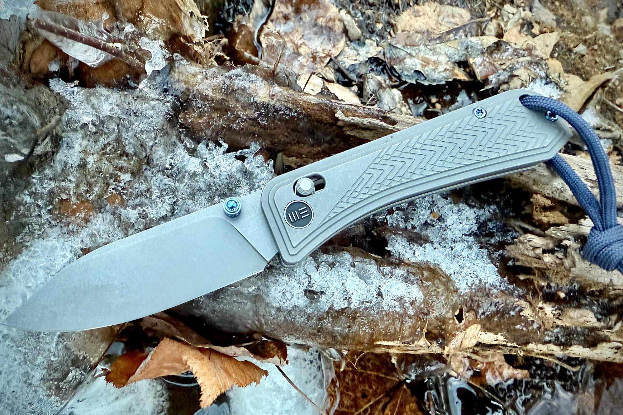 The WE Knife Bougie Banter rests on a frosted log, showcasing its sleek design and textured handle against a natural background