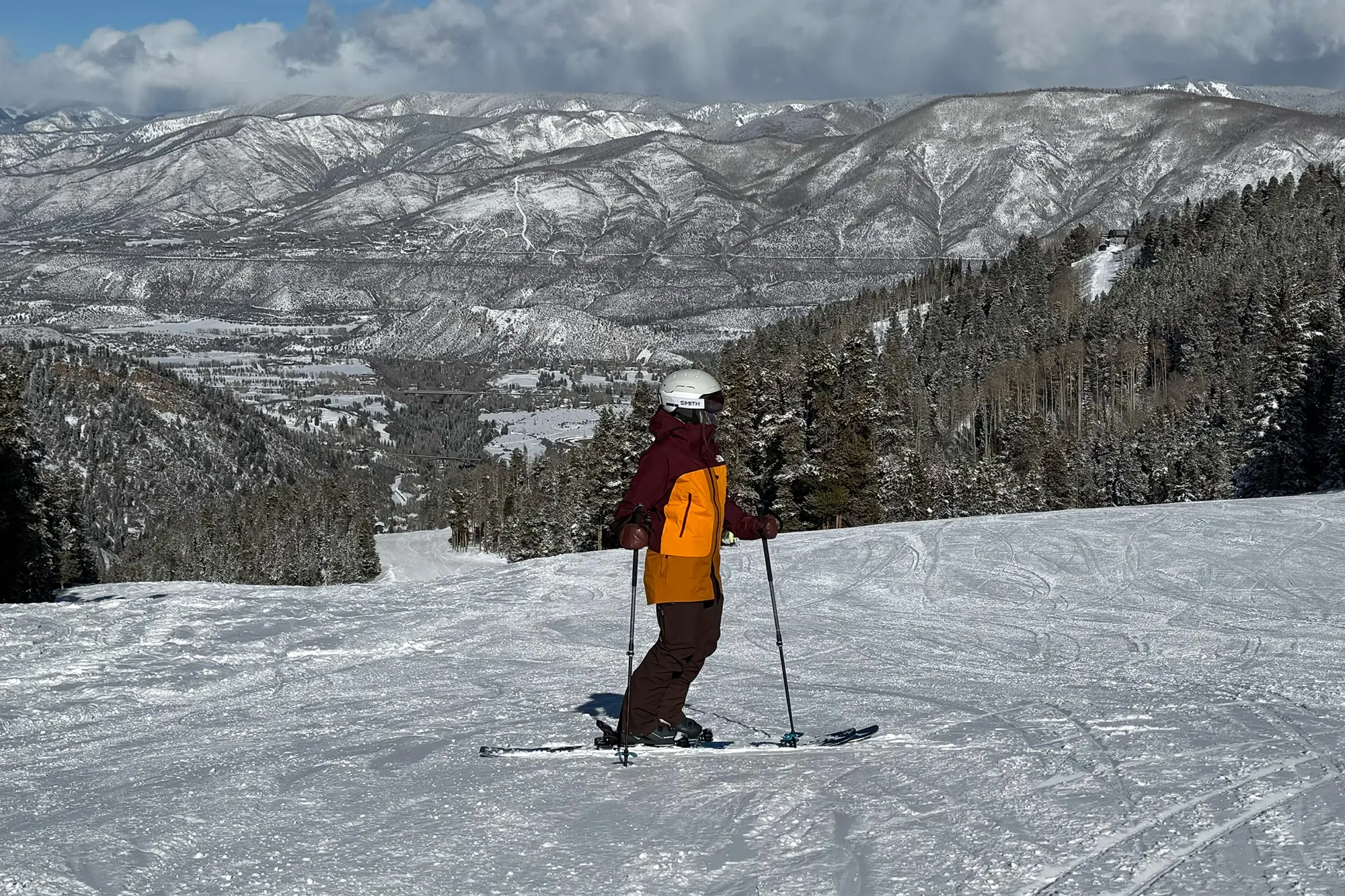A skier dressed in The North Face Summit Series Verbier Gore-Tex Jacket pauses mid-run, set against a backdrop of snow-covered mountains and a scenic valley below
