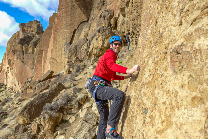 A climber in a red jacket and Outdoor Research Ferrosi pants ascends a vertical rock face with climbing gear and a blue helmet